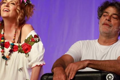 Dois artistas em um palco, uma mulher sorrindo e um homem com expressão relaxada.