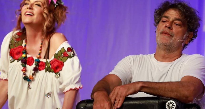 Dois artistas em um palco, uma mulher sorrindo e um homem com expressão relaxada.