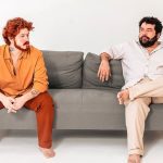 Dois homens sentados em um sofá cinza, um com roupa laranja e o outro com roupa clara.