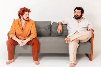 Dois homens sentados em um sofá cinza, um com roupa laranja e o outro com roupa clara.