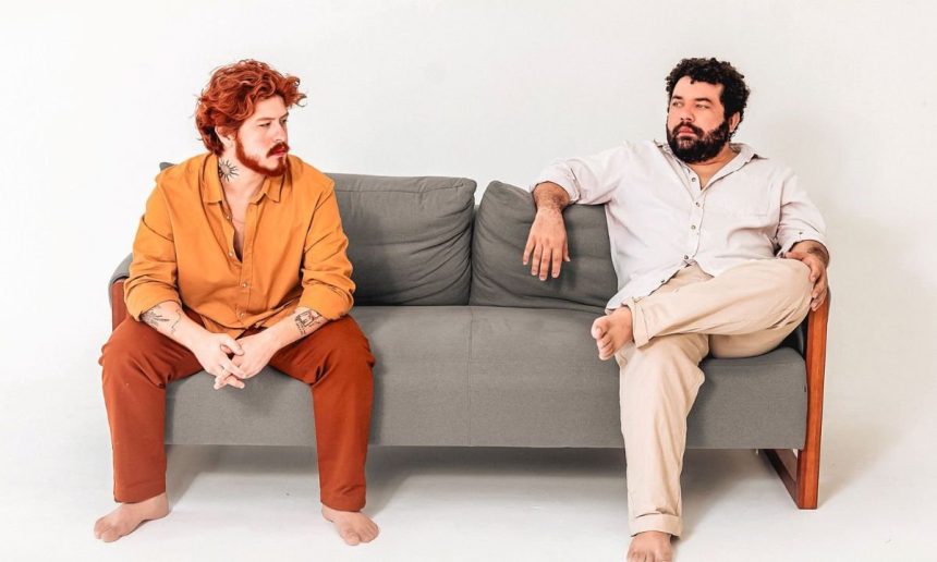 Dois homens sentados em um sofá cinza, um com roupa laranja e o outro com roupa clara.