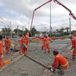 Equipe de construção civil trabalhando em obra de asfalto, usando equipamentos e uniformes laranja.