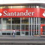 Fachada da agência Santander, com logotipo e letreiro em vermelho e branco, em um dia ensolarado.
