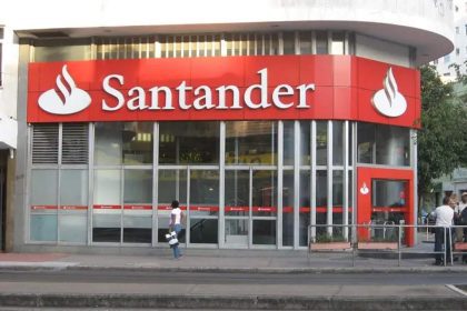 Fachada da agência Santander, com logotipo e letreiro em vermelho e branco, em um dia ensolarado.