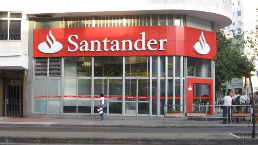 Fachada da agência Santander, com logotipo e letreiro em vermelho e branco, em um dia ensolarado.