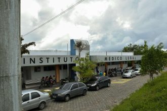 Fachada do Instituto Médico Legal com carros estacionados e pessoas, em um dia nublado.