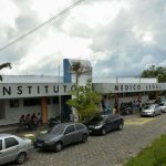 Fachada do Instituto Médico Legal (IML) com carros estacionados e pessoas na frente.
