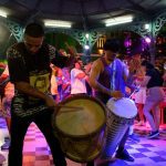 Grupo tocando tambores e instrumentos de sopro em apresentação ao ar livre, com luzes coloridas.