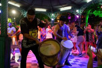 Grupo tocando tambores e instrumentos de sopro em apresentação ao ar livre, com luzes coloridas.
