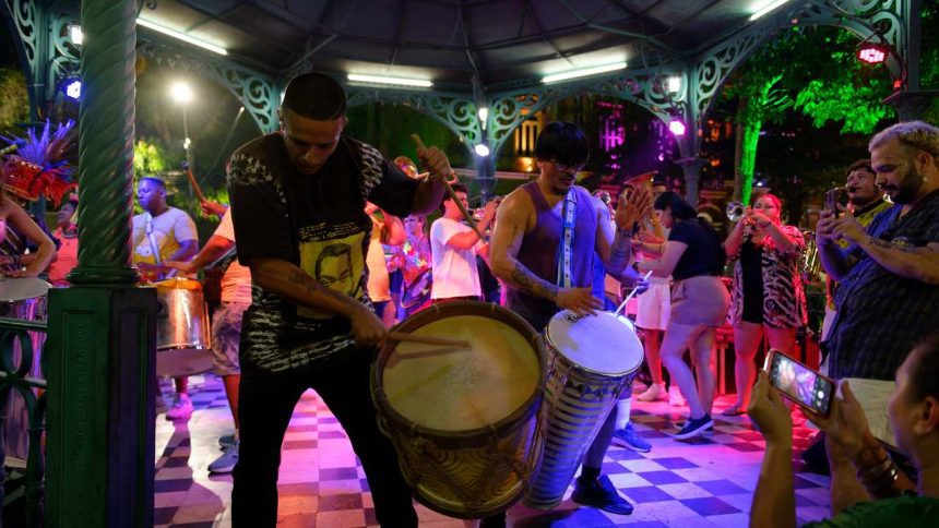 Grupo tocando tambores e instrumentos de sopro em apresentação ao ar livre, com luzes coloridas.
