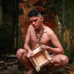 Homem agachado com colar e banquinho, em cenário de ruínas, parte do espetáculo 'A Ilha Profana do Cantagalo'.