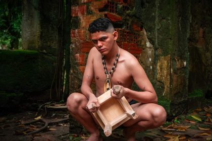 Homem agachado com colar e banquinho, em cenário de ruínas, parte do espetáculo 'A Ilha Profana do Cantagalo'.
