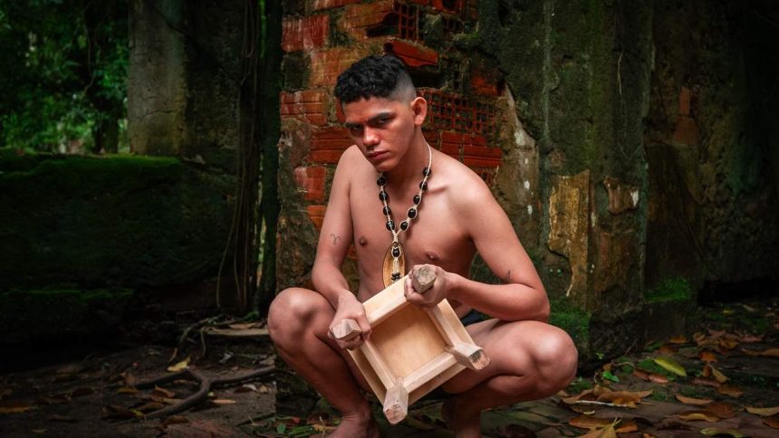 Homem agachado com colar e banquinho, em cenário de ruínas, parte do espetáculo 'A Ilha Profana do Cantagalo'.