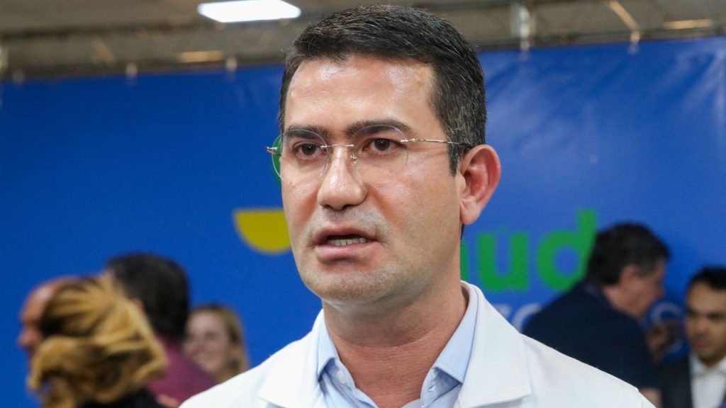 Homem com óculos fala em evento, com fundo azul e pessoas ao fundo.