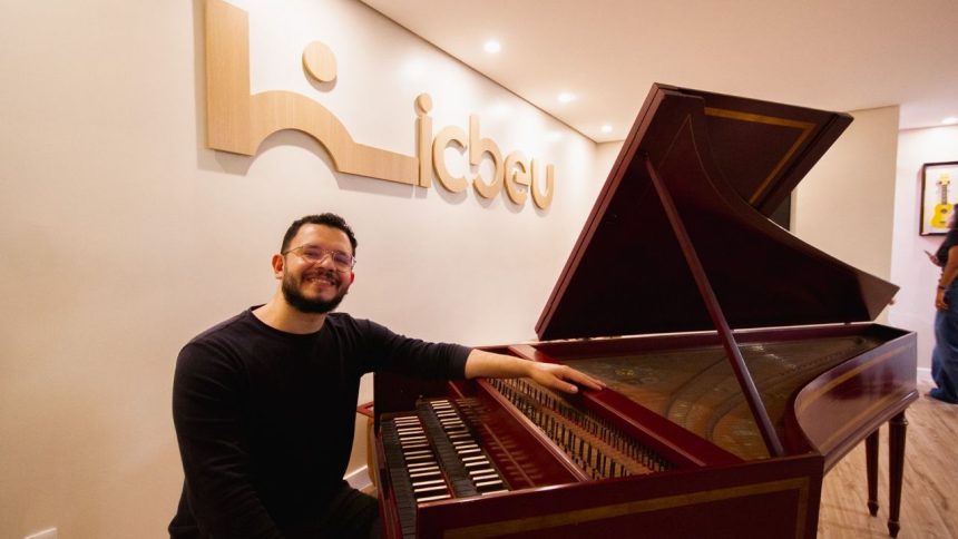 Homem sorrindo ao lado de cravo, instrumento musical barroco, no ICBEU Manaus.