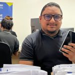 Homem sorrindo segura celular, em escritório com banner da PolÃcia Amazonas e sistema de recuperação de celulares.