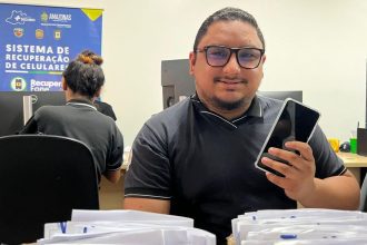 Homem sorrindo segura celular, em escritório com banner da Polícia Amazonas e sistema de recuperação de celulares.