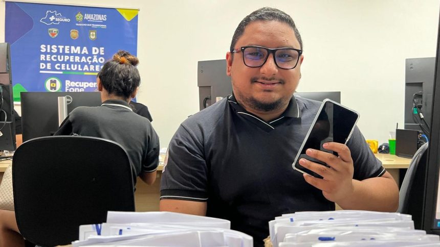 Homem sorrindo segura celular, em escritório com banner da Polícia Amazonas e sistema de recuperação de celulares.