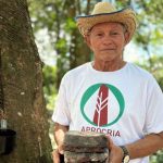 Homem sorrindo segurando borracha de árvore, ao lado de tronco de árvore com cortes para extração de látex.