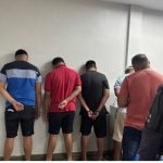 Homens detidos em uma delegacia, com as mãos para trás.