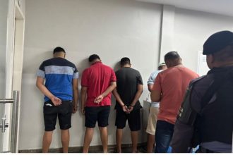 Homens detidos em uma delegacia, com as mãos para trás.