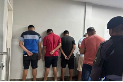 Homens detidos em uma delegacia, com as mãos para trás.