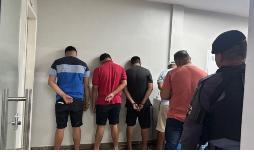 Homens detidos em uma delegacia, com as mãos para trás.