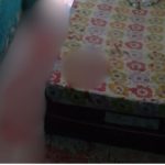 Imagem borrada de um homem próximo a uma cama com lençóis floridos, possivelmente em um ambiente interno.