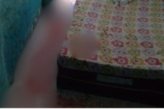 Imagem borrada de um homem próximo a uma cama com lençóis floridos, possivelmente em um ambiente interno.