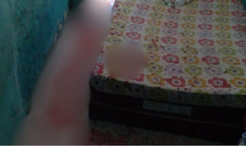 Imagem borrada de um homem próximo a uma cama com lençóis floridos, possivelmente em um ambiente interno.