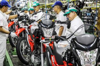 Linha de montagem de motocicletas Honda em fábrica, trabalhadores uniformizados inspecionando motos.