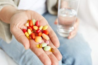 Mão com comprimidos coloridos e um copo de água.