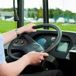 Motorista de ônibus segurando o volante, com painel e paisagem ao fundo.