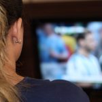 Mulher assistindo jogo na TV, com destaque para a orelha com brincos e cabelo loiro.
