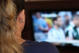 Mulher assistindo jogo na TV, com destaque para a orelha com brincos e cabelo loiro.