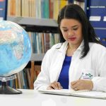 Mulher escrevendo em caderno com globo terrestre e estante de livros ao fundo.