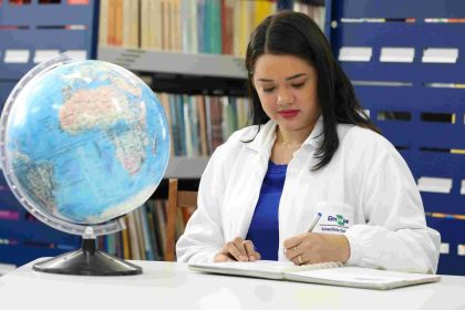 Mulher escrevendo em caderno com globo terrestre e estante de livros ao fundo.