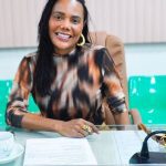 Mulher sorrindo em uma reunião, usando blusa estampada e brincos dourados.