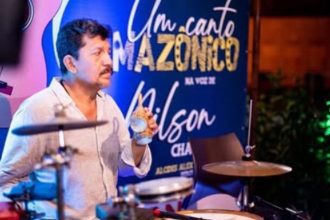 Músico com copo na mão em show. Ao fundo, banner 'Um Canto Amazônico'.