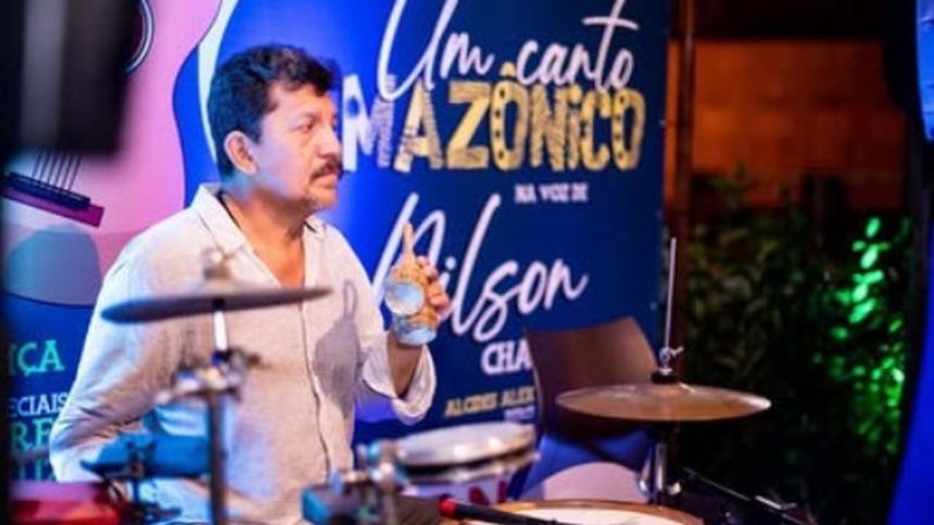 Músico com copo na mão em show. Ao fundo, banner 'Um Canto Amazônico'.