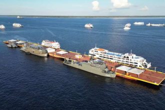 Navios atracados em uma plataforma no rio, com outros navios ao fundo.