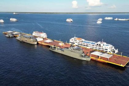 Navios atracados em uma plataforma no rio, com outros navios ao fundo.