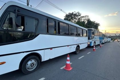 Ônibus brancos estacionados em fila em uma rua, com cones de sinalização e céu ao fundo.