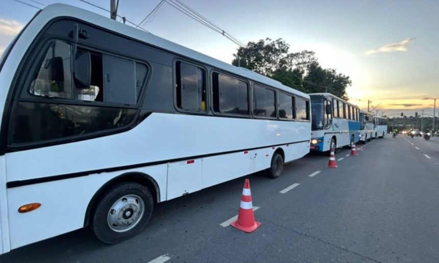 Ônibus brancos estacionados em fila em uma rua, com cones de sinalização e céu ao fundo.