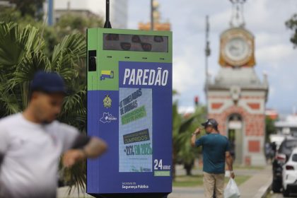 Painel 'PAREDÃO' com nova etapa de reconhecimento facial exibe informações de segurança pública em área urbana, com texto sobre apreensão de drogas e mandados de prisão.