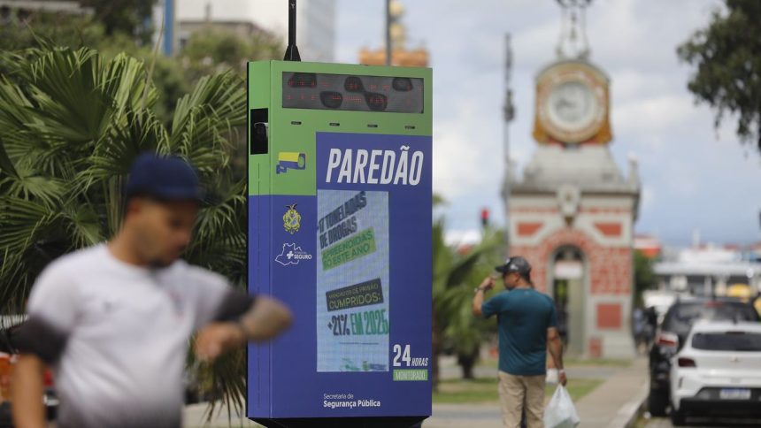 Painel 'PAREDÃO' com nova etapa de reconhecimento facial exibe informações de segurança pública em área urbana, com texto sobre apreensão de drogas e mandados de prisão.