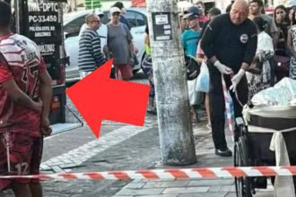 Polícia investiga cena do crime com homem morto em cadeira de rodas, com viatura e pessoas ao redor.