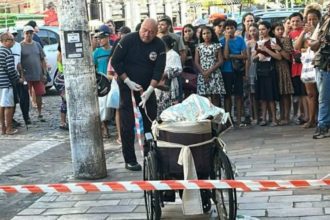 Policial em cena de crime no Centro de Manaus, cadeira de rodas e multidão observando.