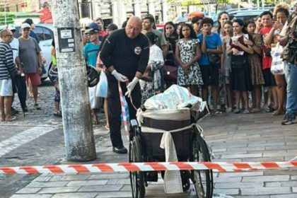 Policial em cena de crime no Centro de Manaus, cadeira de rodas e multidão observando.