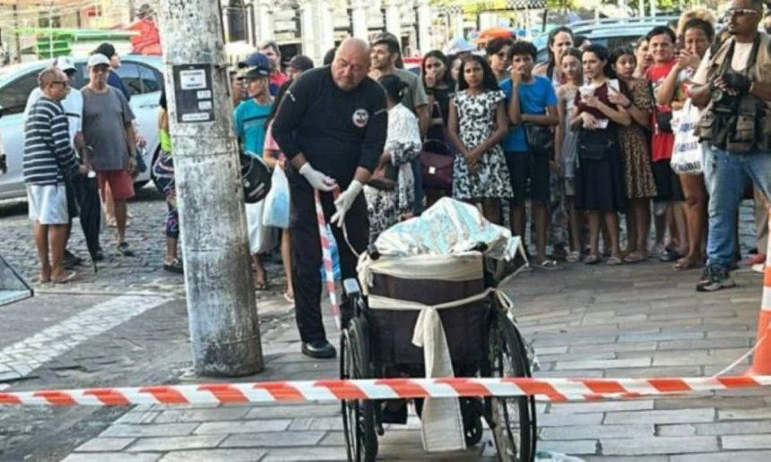 Policial em cena de crime no Centro de Manaus, cadeira de rodas e multidão observando.